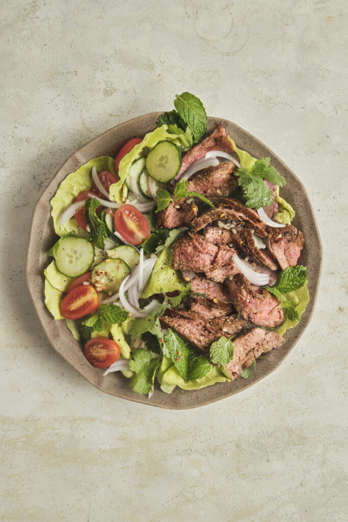 Close-up of Thai-inspired steak salad with sliced medium-rare beef, fresh herbs, cucumber, cherry tomatoes, and lime dressing served over lettuce.