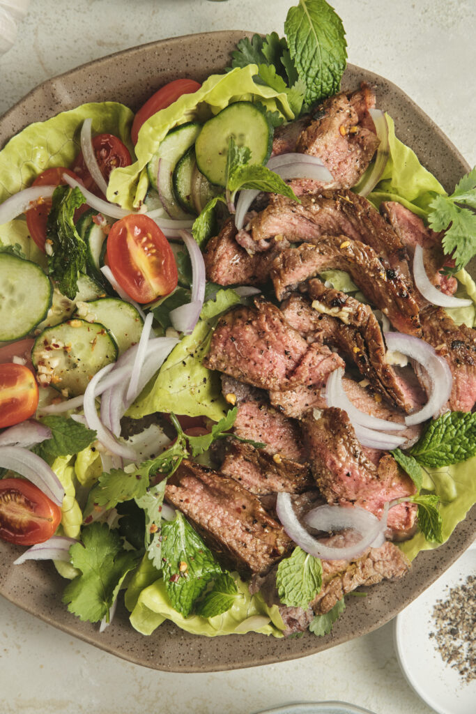 Close-up of Thai-inspired steak salad with sliced medium-rare beef, fresh herbs, cucumber, cherry tomatoes, and lime dressing served over lettuce.