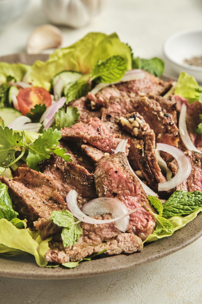 Close-up of Thai-inspired steak salad with sliced medium-rare beef, fresh herbs, cucumber, cherry tomatoes, and lime dressing served over lettuce.