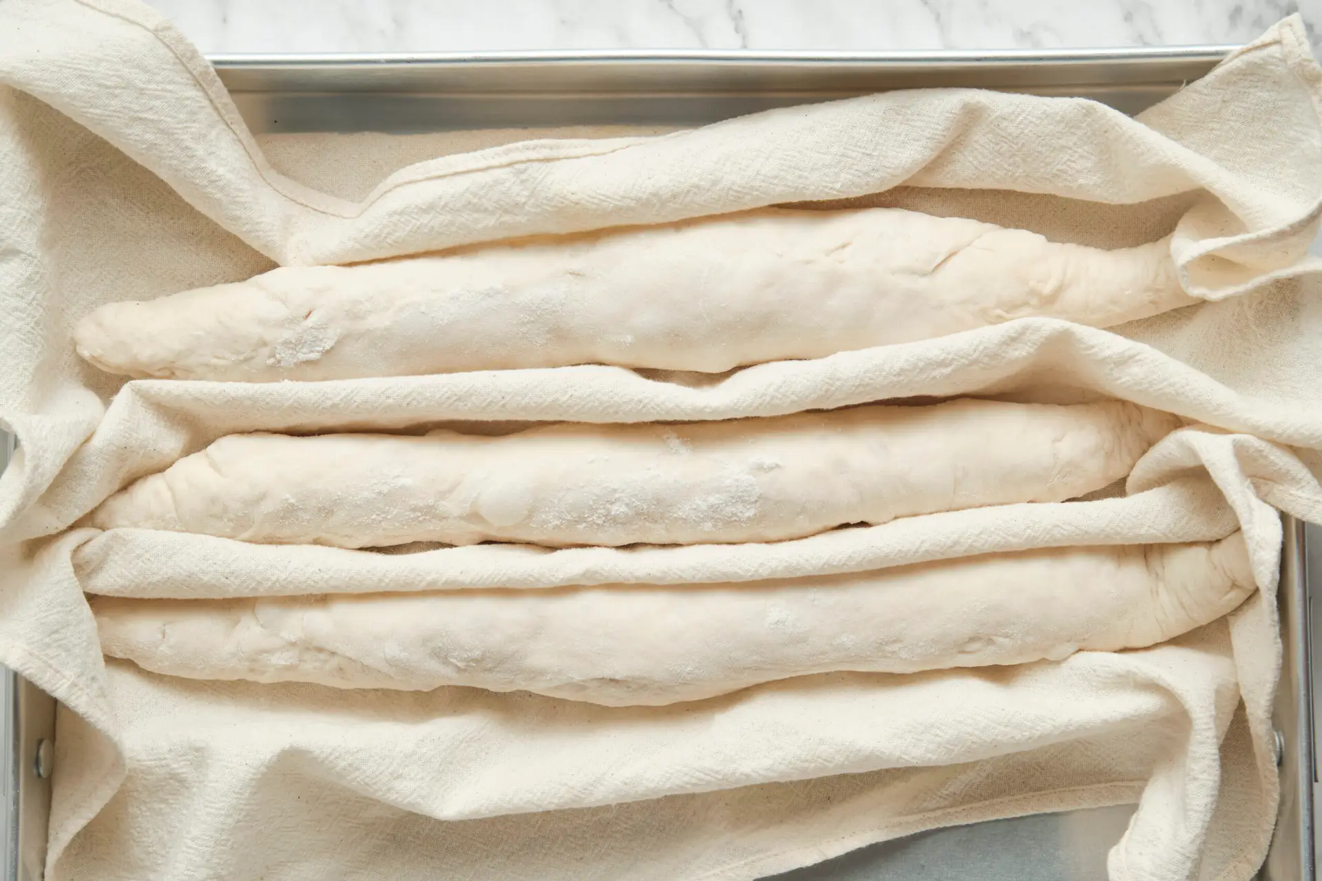 Shaped French baguettes proofing on a cloth before baking—final rise stage in a baguette recipe and how to make baguette at home for a crisp crust and airy crumb.