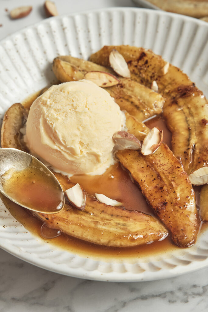 Bananas Foster served on a white plate with scoops of vanilla ice cream, topped with warm caramelized banana rum sauce being spooned over the dessert.