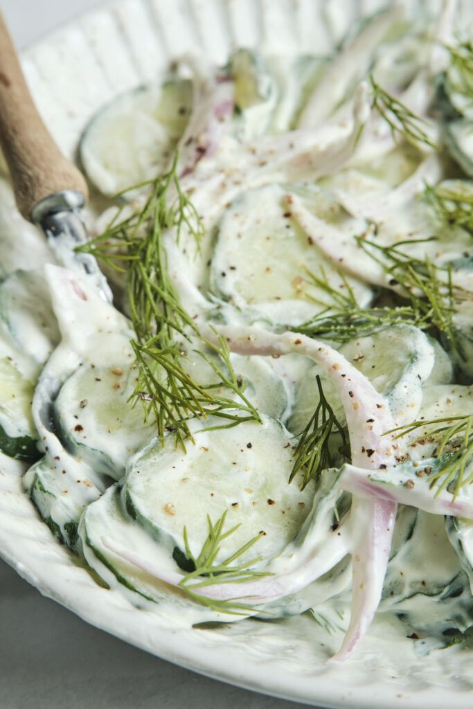 A close-up shot of creamy cucumber salad with dill and red onions in a white bowl, showing a healthy summer side dish.