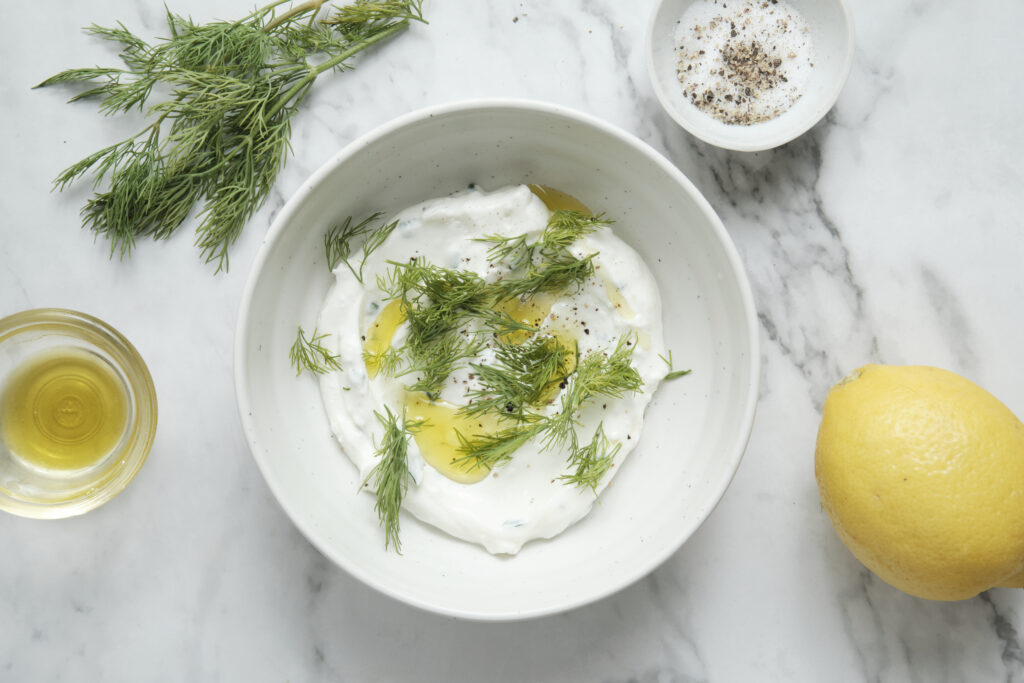 Making the creamy yogurt dressing for a cucumber salad in a white bowl with fresh dill, olive oil, and lemon.