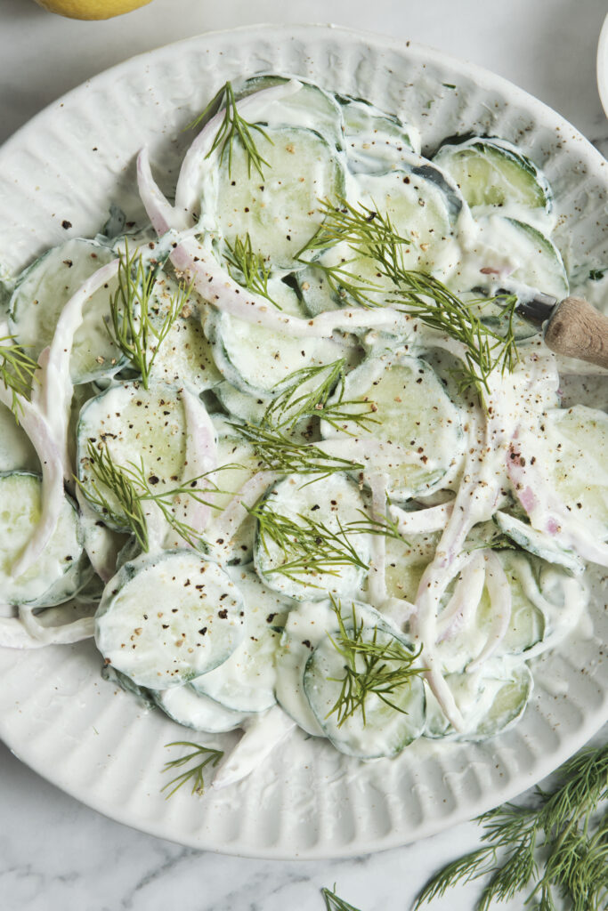 A close-up shot of creamy cucumber salad with dill and red onions in a white bowl, showing a healthy summer side dish.