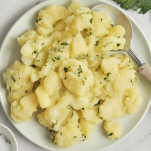 Close-up of German potato salad served in a rustic bowl with sliced potatoes, fresh herbs, red onion, and a light tangy dressing, garnished with parsley and black pepper.
