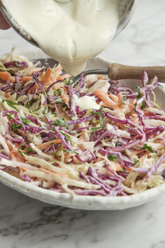 Close-up of creamy Greek yogurt coleslaw with shredded cabbage and carrots as the tangy yogurt dressing is poured over the top, creating a fresh and healthy summer side dish.