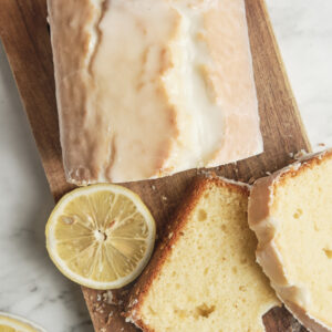 Close-up of a glazed lemon loaf cake sliced on a rustic wooden board, showing the soft and moist texture with fresh lemon slices and crumbs scattered around.