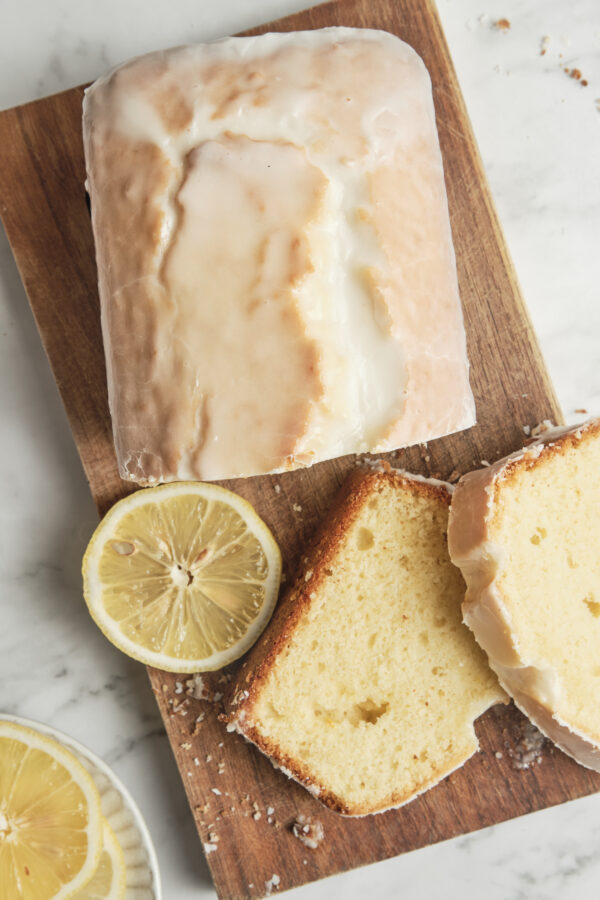 Close-up of a glazed lemon loaf cake sliced on a rustic wooden board, showing the soft and moist texture with fresh lemon slices and crumbs scattered around.