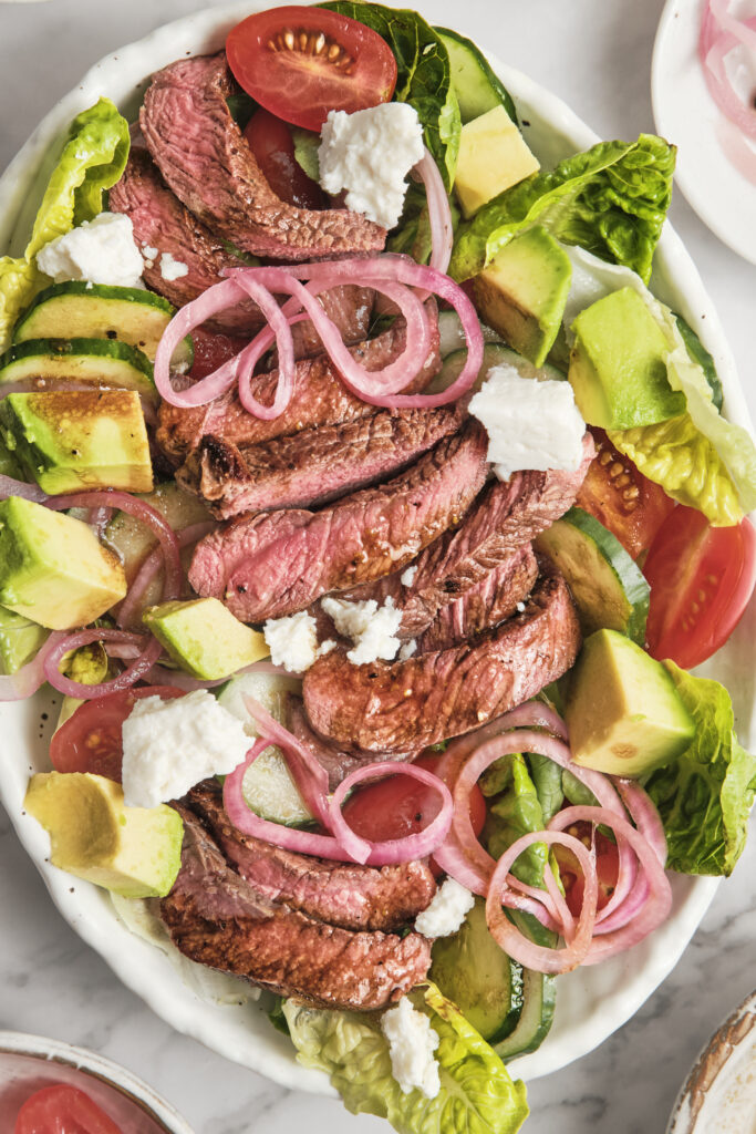 Juicy grilled steak served over fresh greens with avocado, cherry tomatoes, red onions, and creamy balsamic dressing in a healthy high-protein steak salad.