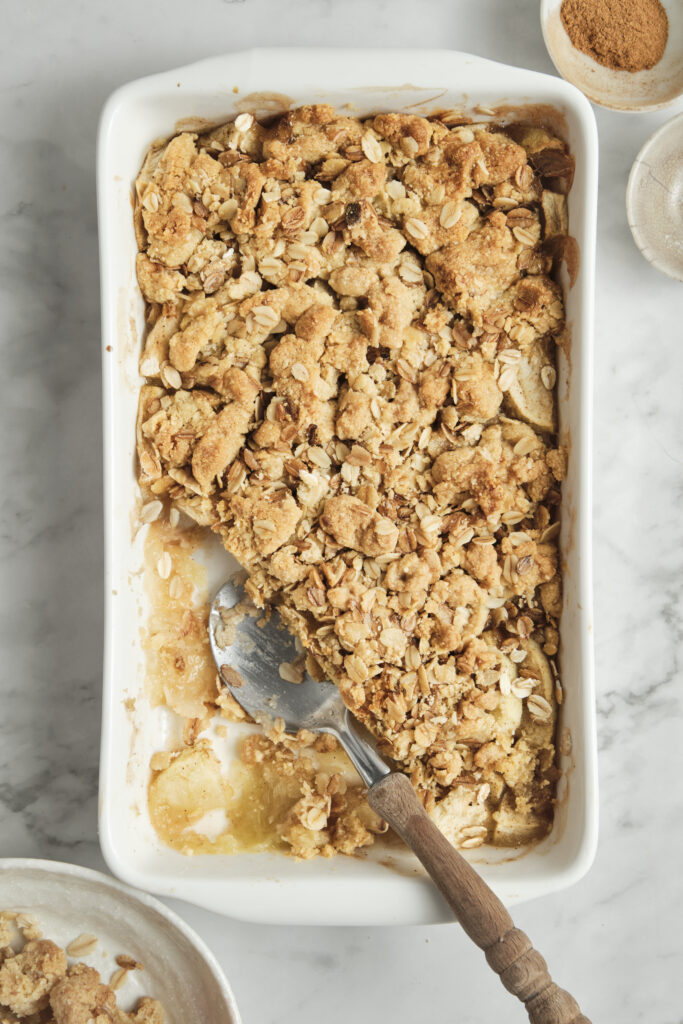 A top-down view of the freshly baked, golden-brown apple crisp in a ceramic baking dish, with a large spoon scooping out a warm serving.