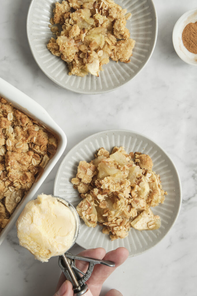 A hand placing a scoop of vanilla ice cream onto a warm serving of apple crisp on a white plate.