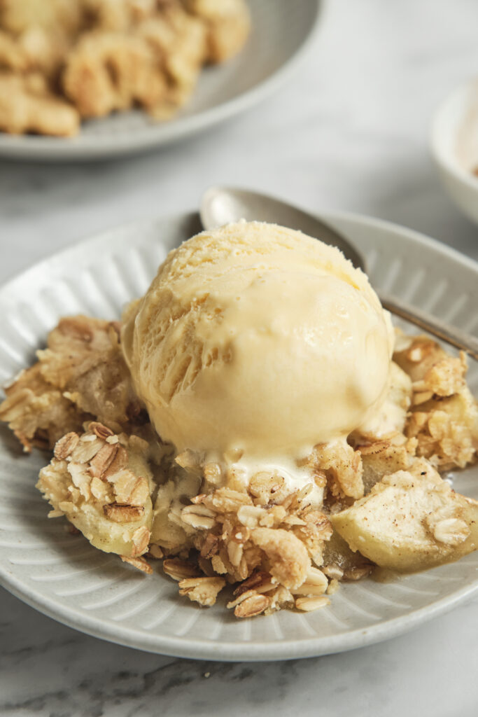 A close-up view of a warm serving of apple crisp in a ceramic bowl, featuring a golden-brown oat and almond flour crumble topping over baked cinnamon apples. A single, melting scoop of vanilla ice cream sits perfectly on top