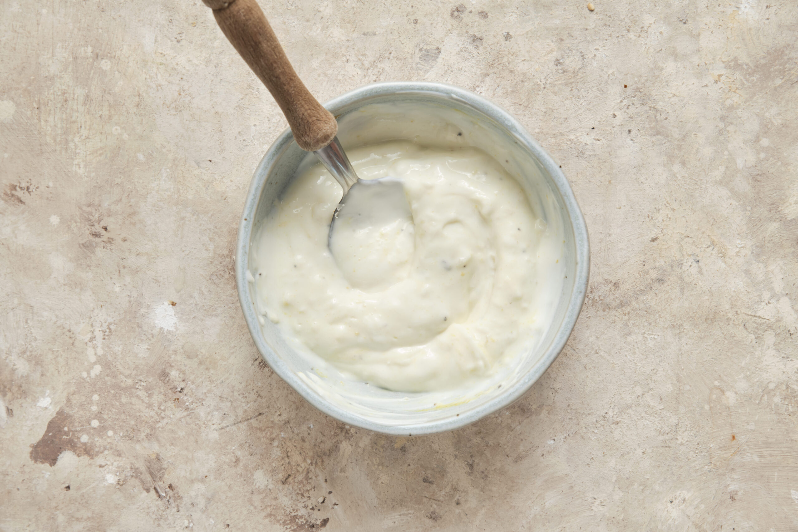 Whisking creamy homemade Caesar dressing ingredients together in a glass bowl with a small spoon.