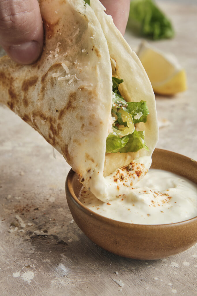 A crispy chicken Caesar smash taco being dipped into a bowl of creamy homemade Caesar dressing.