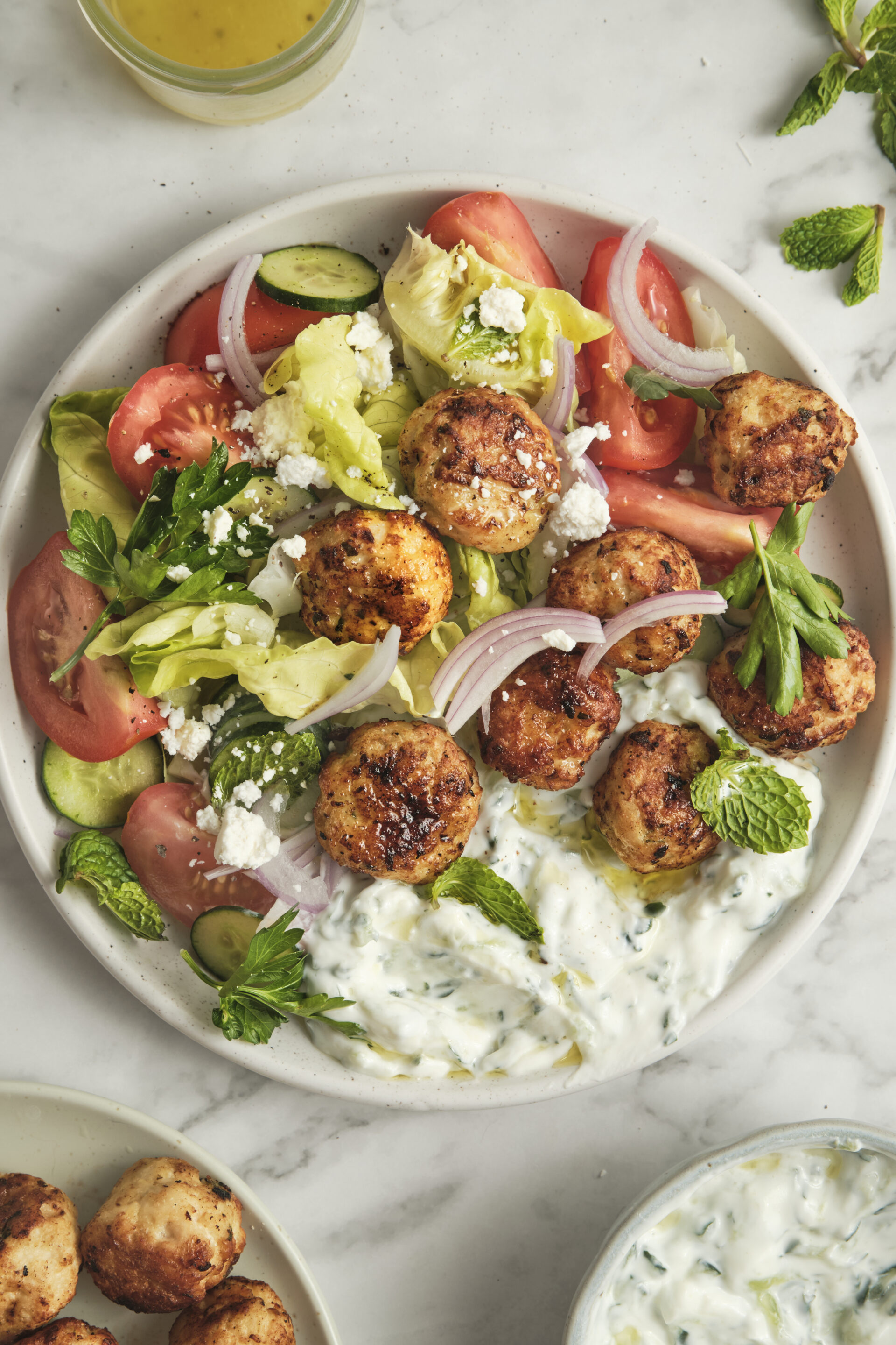 A vibrant Greek chicken bowl filled with baked chicken meatballs, a scoop of creamy homemade tzatziki sauce, crisp cucumbers, juicy tomatoes, sliced red onions, and fresh summer salad greens.