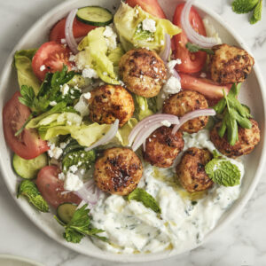 A vibrant Greek chicken bowl filled with baked chicken meatballs, a scoop of creamy homemade tzatziki sauce, crisp cucumbers, juicy tomatoes, sliced red onions, and fresh summer salad greens.