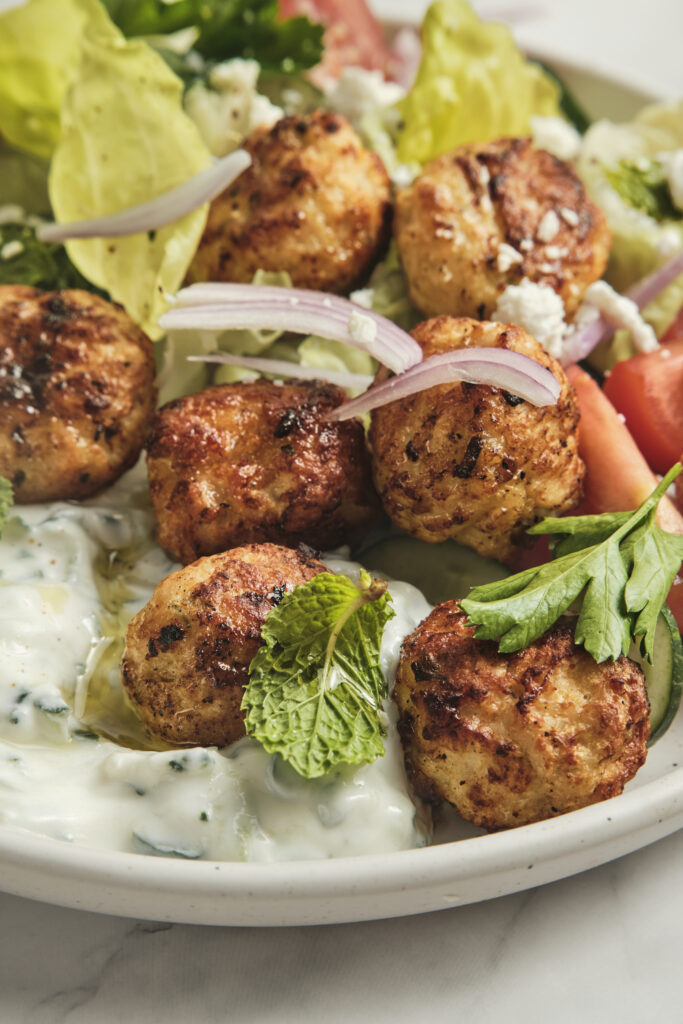 A detailed close-up of a juicy, golden-brown baked chicken meatball being dipped into creamy, homemade tzatziki sauce with fresh dill.