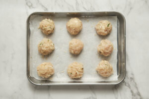 Placing evenly portioned chicken meatballs onto a parchment-lined baking sheet using a cookie dough scoop.