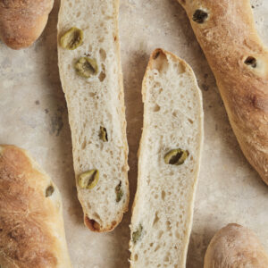A close-up shot of an easy olive baguette sliced open, revealing a fluffy, airy interior crumb packed with chopped green olives and a perfectly golden, crispy crust.