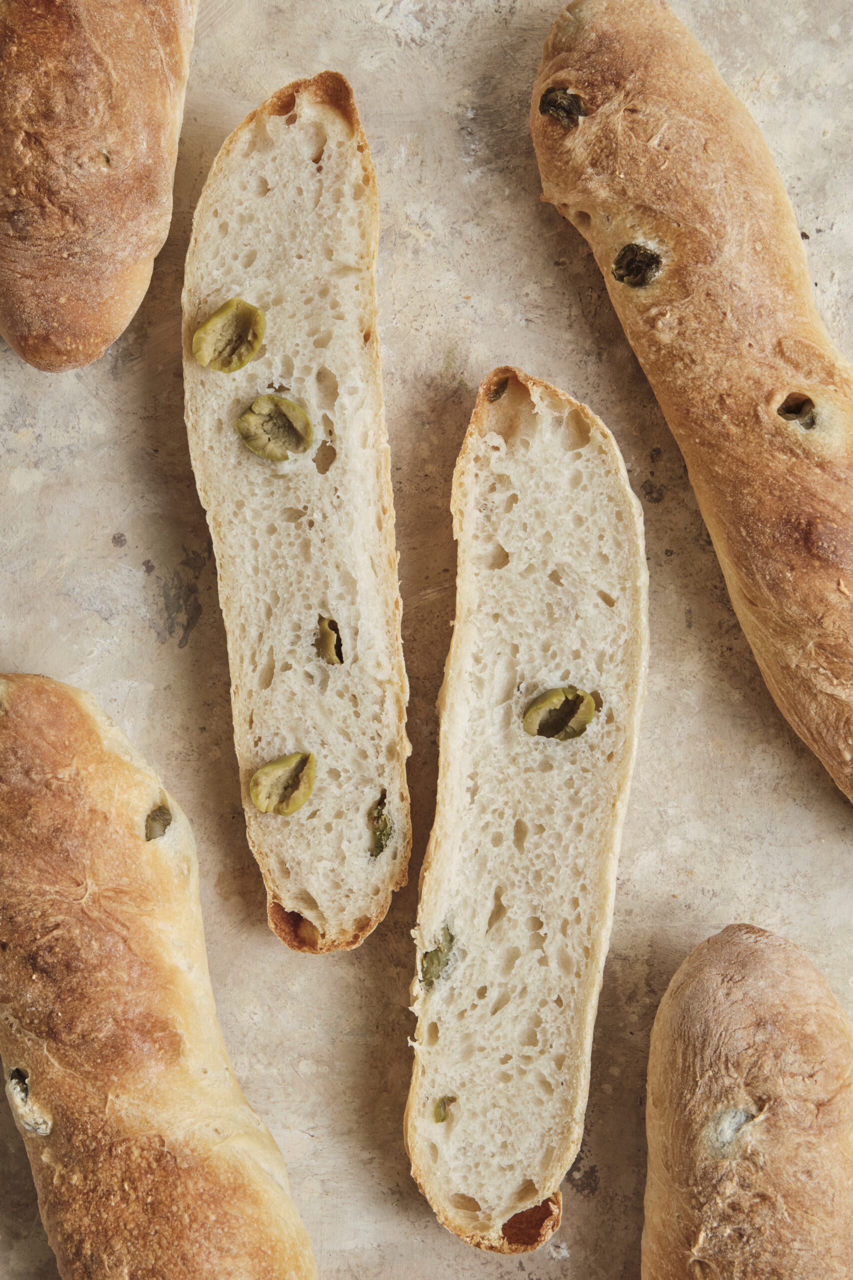 A close-up shot of an easy olive baguette sliced open, revealing a fluffy, airy interior crumb packed with chopped green olives and a perfectly golden, crispy crust.