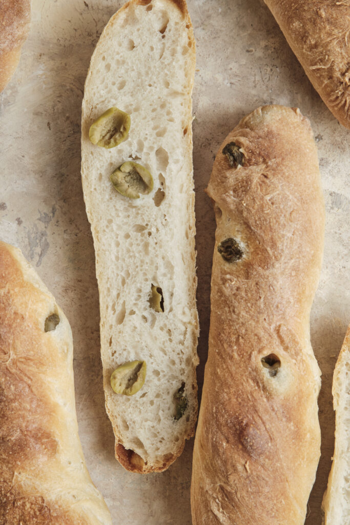 A close-up shot of an easy olive baguette sliced open, revealing a fluffy, airy interior crumb packed with chopped green olives and a perfectly golden, crispy crust.