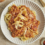 Hearty vegan lentil bolognese sauce served over a bed of wide tagliatelle pasta in a deep plate, set against a rustic background.