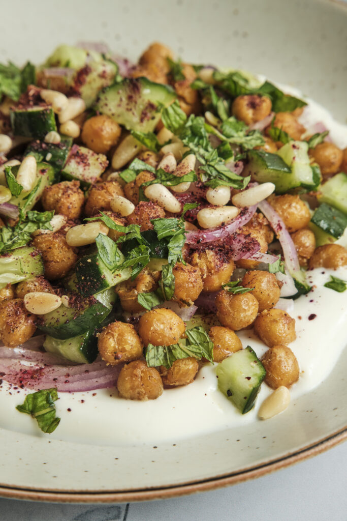 A refreshing roasted chickpea and cucumber salad with fresh herbs and a creamy tahini dressing, beautifully served on a plate and topped with toasted pine nuts.