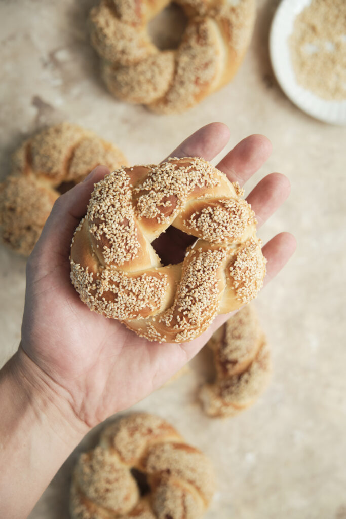 A close-up of a person holding a freshly baked, golden-brown Turkish simit heavily coated in toasted sesame seeds.