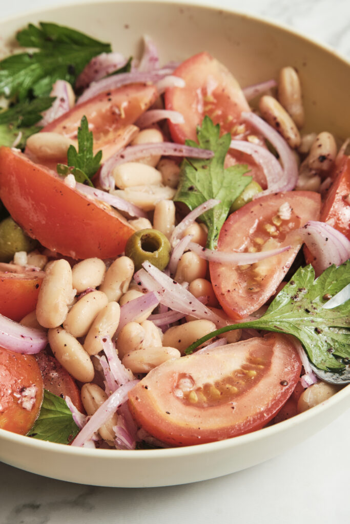 Close-up shot of a Mediterranean white bean salad with vibrant red tomatoes, green olives, and fresh minced herbs.