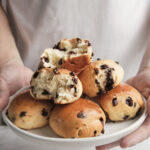 Hands holding freshly baked chocolate brioche buns arranged on a plate, showing their soft, fluffy texture and rich chocolate filling.