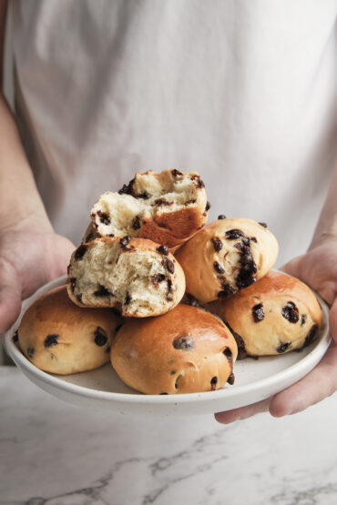 Hands holding freshly baked chocolate brioche buns arranged on a plate, showing their soft, fluffy texture and rich chocolate filling.