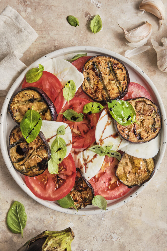 An Italian eggplant Caprese made with layers of roasted eggplant, fresh mozzarella, ripe tomatoes, and basil, finished with a drizzle of balsamic glaze and served on a rustic ceramic plate.