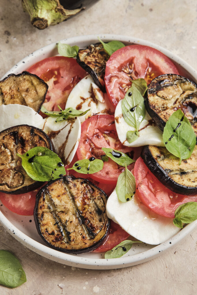 An Italian eggplant Caprese made with layers of roasted eggplant, fresh mozzarella, ripe tomatoes, and basil, finished with a drizzle of balsamic glaze and served on a rustic ceramic plate.